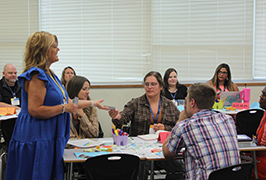  A woman addresses a room of other seated adults and hands one a grey card